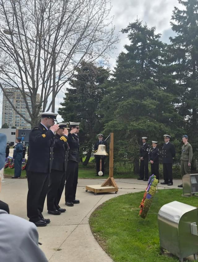 Uniformed naval personnel salute beside a ship's bell on a wooden stand, with a wreath on the grass and others standing at attention.