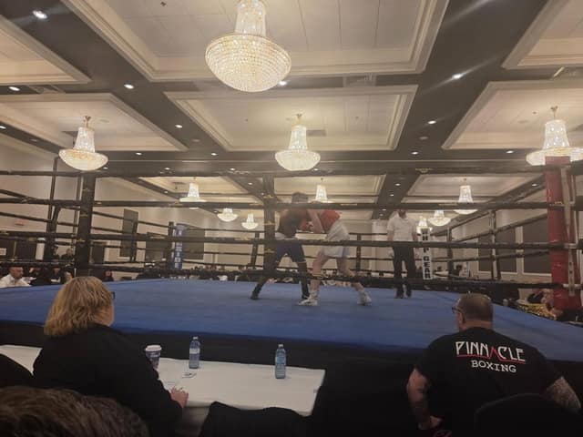 Two boxers trade punches in a blue ring while a referee watches and spectators sit at a table in the foreground.