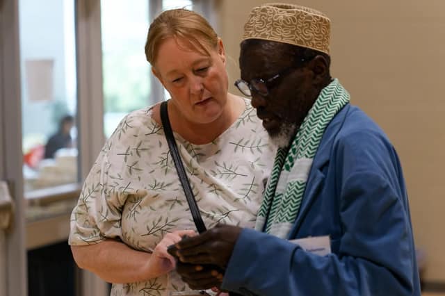 A woman in a leaf-patterned blouse looks at a phone held by an older man in a cap, glasses, green scarf and blue jacket.