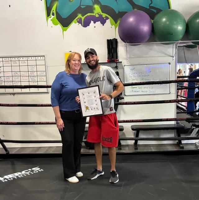 A woman in a blue top and a bearded man in a cap and red shorts hold a framed certificate together beside a boxing ring.