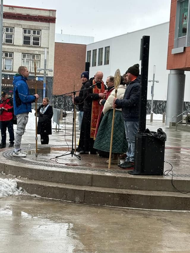 A priest in red and gold vestments speaks into a microphone on a stepped platform in the snow, with men holding long staffs beside him.