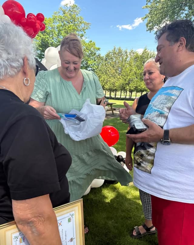 A woman in a green dress laughs while opening a gift bag, with a man in a white shirt and others nearby among balloons.