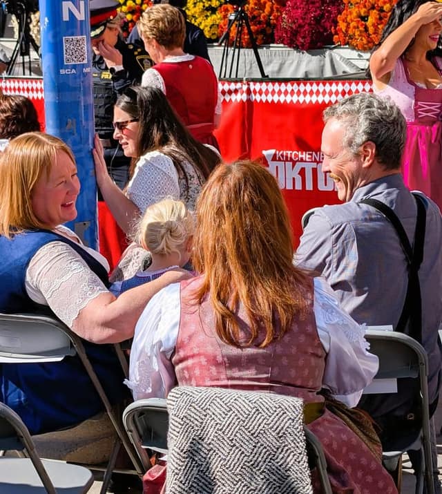 A woman, a man and a young girl sit in folding chairs, laughing, near a red Oktoberfest stage skirt and flower displays.