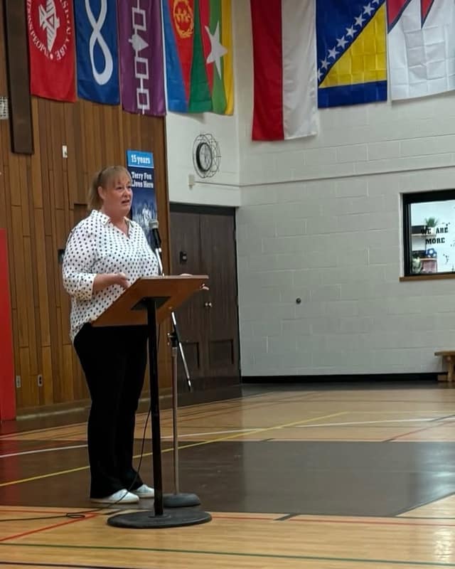 A woman in a polka-dot blouse speaks at a wooden lectern in a school gym, with flags and a Terry Fox banner on the wall behind her.