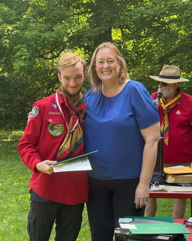 A smiling woman in a blue top stands beside a young man in a red Scouts uniform holding a certificate, with an older scouter behind.