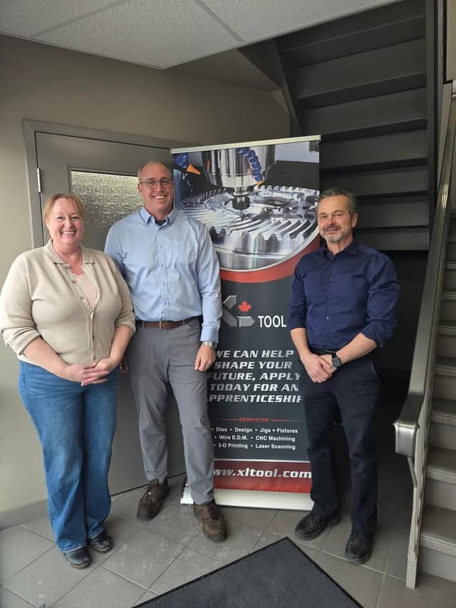 Three people stand smiling beside an XL Tool banner about apprenticeships and services, in a lobby near a staircase.