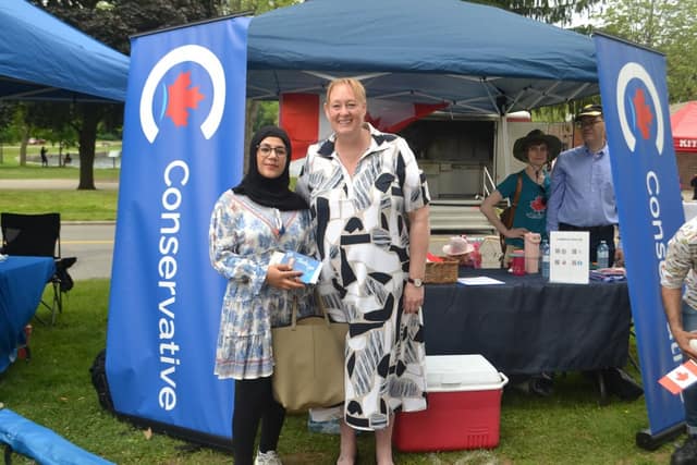 Kelly DeRidder stands with a woman in a black hijab between blue Conservative banners at a festival tent with a table and Canadian flags.