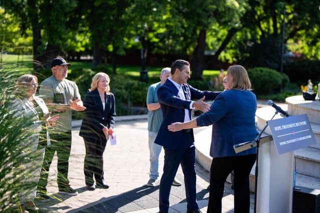 A man in a navy suit extends his arms to greet a woman at a podium as onlookers stand nearby in a sunny park plaza.