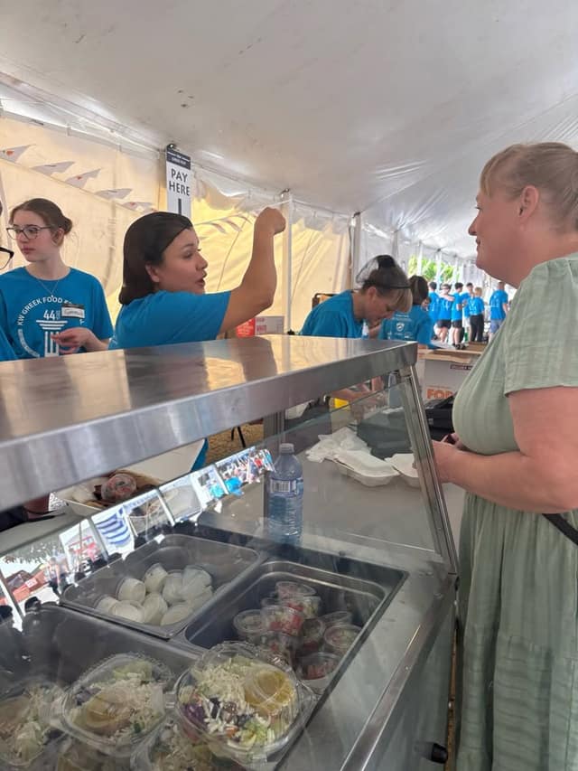 A woman in a green top talks with a volunteer in a blue shirt at a food counter under a tent with a 'Pay Here' sign.