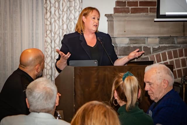 A woman in a dark blazer speaks at a wooden podium with her hands open while seated guests listen in a room with a brick fireplace.