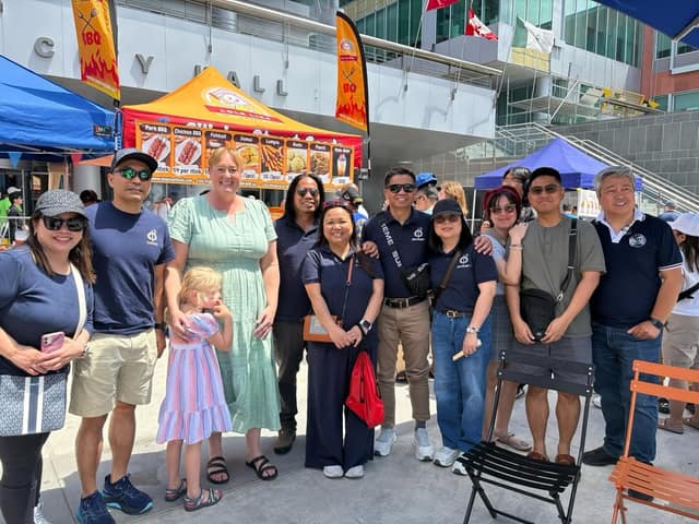 A woman in a green dress and a young girl pose with a group of smiling adults in front of a BBQ food tent outside City Hall.
