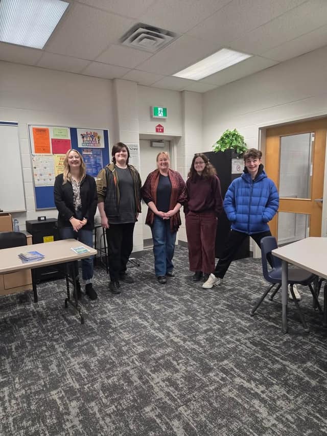 A woman in a red shawl stands in a classroom with four other people, including students, near a bulletin board and a door.