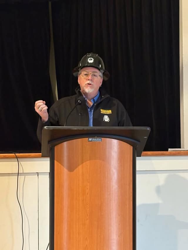 A man in a black hard hat and jacket speaks at a wooden podium with a microphone, one fist raised, before a dark curtain.
