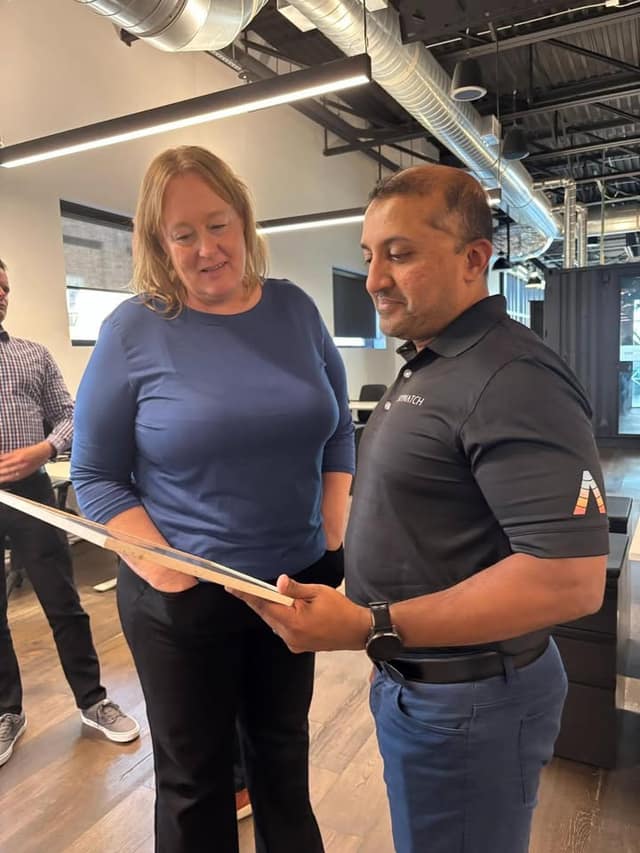 A woman in a blue top and a man in a black SkyWatch polo look down at a framed document in an open office with exposed ducts.