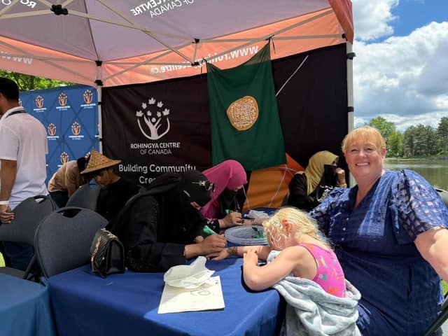 A smiling woman in a blue dress sits at a table under a Rohingya Centre of Canada tent as a young girl watches women do henna designs.