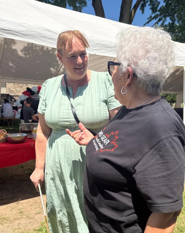 A smiling woman in a green dress talks with a gray-haired woman in a black Rapid Gear shirt under a white tent.
