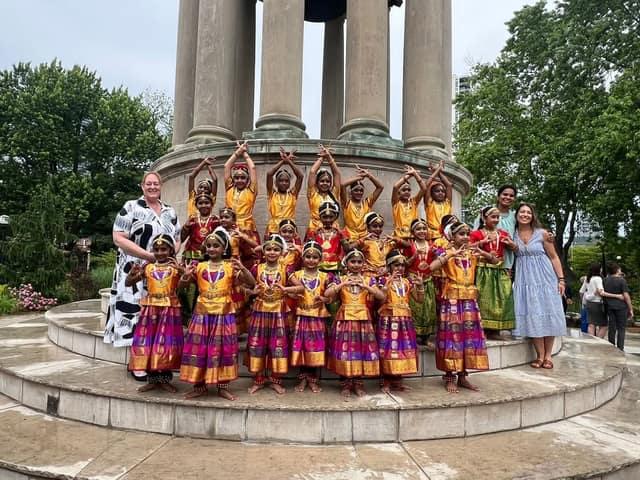 Young dancers in gold, red and purple costumes pose with hands raised in front of a stone monument, with two women standing at the sides.
