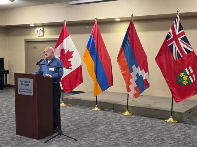 A man in a blue shirt speaks at a podium marked Armenian Community Centre of Cambridge, beside Canadian, Armenian, Artsakh and Ontario flags.