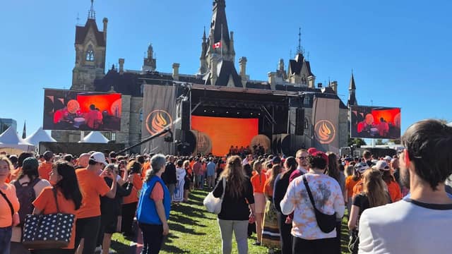 A large crowd in orange shirts faces an outdoor stage with video screens and a flame logo in front of a stone Parliament building.