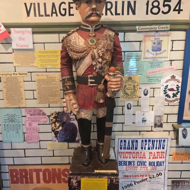 Painted statue of a uniformed officer with a sword beside a "Grand Opening Victoria Park 1896" poster and "Village of Berlin 1854" sign.