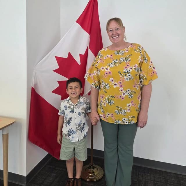 A smiling woman in a yellow floral top holds hands with a young boy in a patterned shirt beside a Canadian flag.