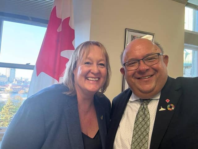 A close-up selfie of a smiling woman and Mayor Berry Vrbanovic, who wears glasses and lapel pins, near a Canadian flag and window.