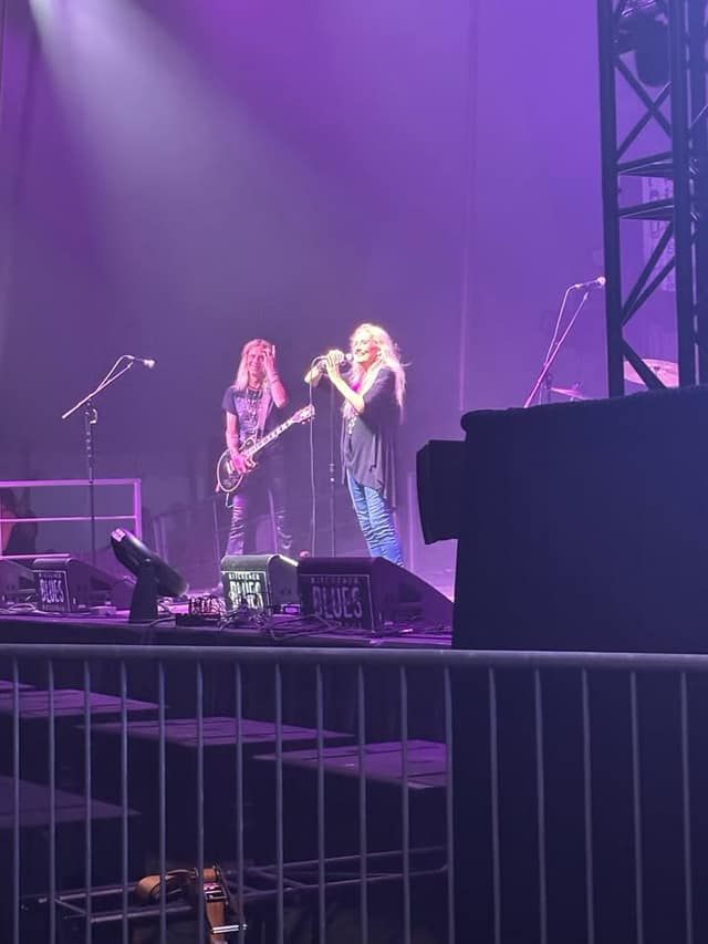 A blonde singer holds a microphone beside a guitarist on stage, with speaker monitors marked Kitchener Blues in front.