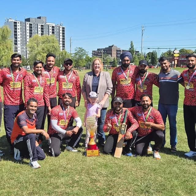 A woman and a small child stand with a cricket team in red patterned jerseys and medals, holding a trophy and a bat on a grassy field.