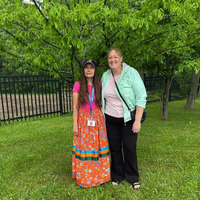 A woman in a mint jacket stands smiling beside a woman in a pink top and orange ribbon skirt under a leafy tree, by a black fence.