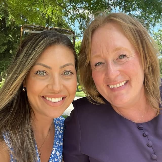 Two smiling women take a close-up selfie outdoors under green trees, one with sunglasses on her head and one in a purple top.