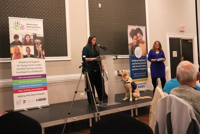 A woman speaks at a podium on a stage as a yellow service dog sits beside her and another woman holds papers near Child Witness Centre banners.