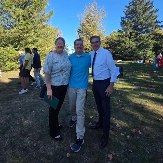 Three people stand arm in arm on a sunny lawn among trees; two are smiling and one holds a green folder, with others behind.
